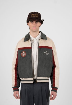 Muž v béžovošedé a červené závodnické bundě přes pruhované tričko a hnědou čepici s nápisem "Umění být pozdě".