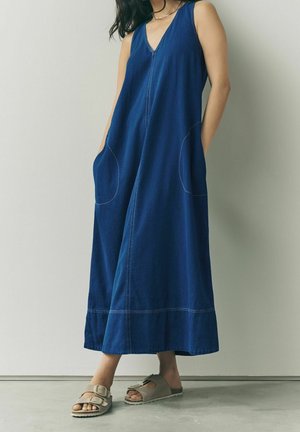 Vrouw in een mouwloze, diepblauwe maxi-jurk met zijzakken en beige sandalen met gesp, staand tegen een eenvoudige lichte muur.