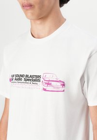 Hvit bomullst-skjorte med et rosa grafikk av en bil og teksten "UF SOUND BLASTERS Car Audio Specialists" over brystet. Korte ermer.
