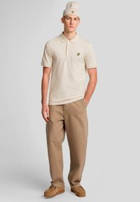 Jonge man met een beige poloshirt, bijpassende muts, beige broek en bruine schoenen, staand met één hand in de zak tegen een effen achtergrond.