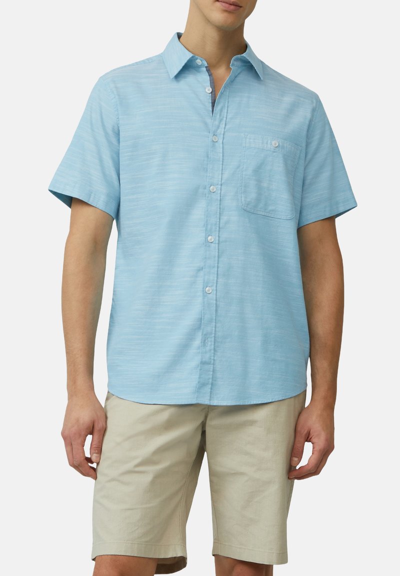 Camicia azzurro chiaro a maniche corte realizzata in tessuto testurizzato, con colletto button-down e taschino sul petto. Abbinata a pantaloni beige.