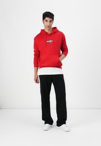Red hooded sweatshirt with a central logo, paired with a white layered shirt and black sweatpants, complemented by white sneakers.