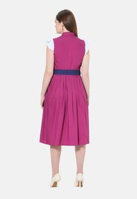 Vestido fucsia con mangas blancas con volantes, cintura ceñida y falda plisada. Presenta un cinturón azul marino y un patrón de lunares.