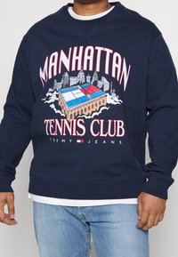 Felpa navy con un grafico di un campo da tennis con edifici, "MANHATTAN TENNIS CLUB" e "TOMMY JEANS" in lettere in grassetto.