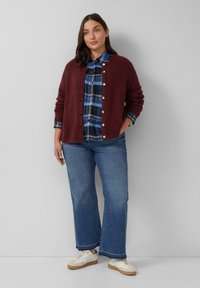 Bordeauxfarbener gestrickter Cardigan mit Knöpfen über einem blauen karierten Hemd, kombiniert mit hellblauen weiten Jeans und weißen Sneakern, in stehender Pose.