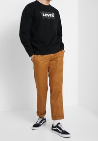 Felpa nera con logo Levi’s camouflage, abbinata a pantaloni beige e sneakers neri. I pantaloni sono arrotolati alle caviglie.