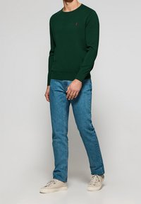 Ciemnozielony sweter z okrągłym dekoltem i ściągaczami na rękawach, w połączeniu z jasnoniebieskimi dżinsami i kremowymi trampkami.