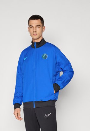 Veste de sport bleue avec un col noir, fermeture éclair à l'avant et poignets élastiques. Présente un logo circulaire en bleu-vert et un petit swoosh Nike sur la poitrine.