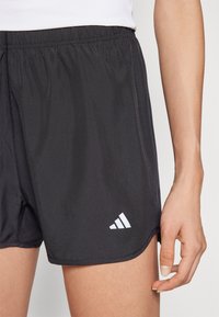 Mustat urheilusortsit, joissa on joustava vyötärö, ja alhaalla vasemmalla puolella valkoinen Adidas-logo sekä sileä, kevyt kangas.