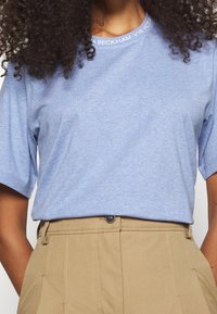 Camiseta de algodón azul claro con mangas cortas, con un escote redondo y texto impreso en blanco. Combinada con pantalones beige de cintura alta.