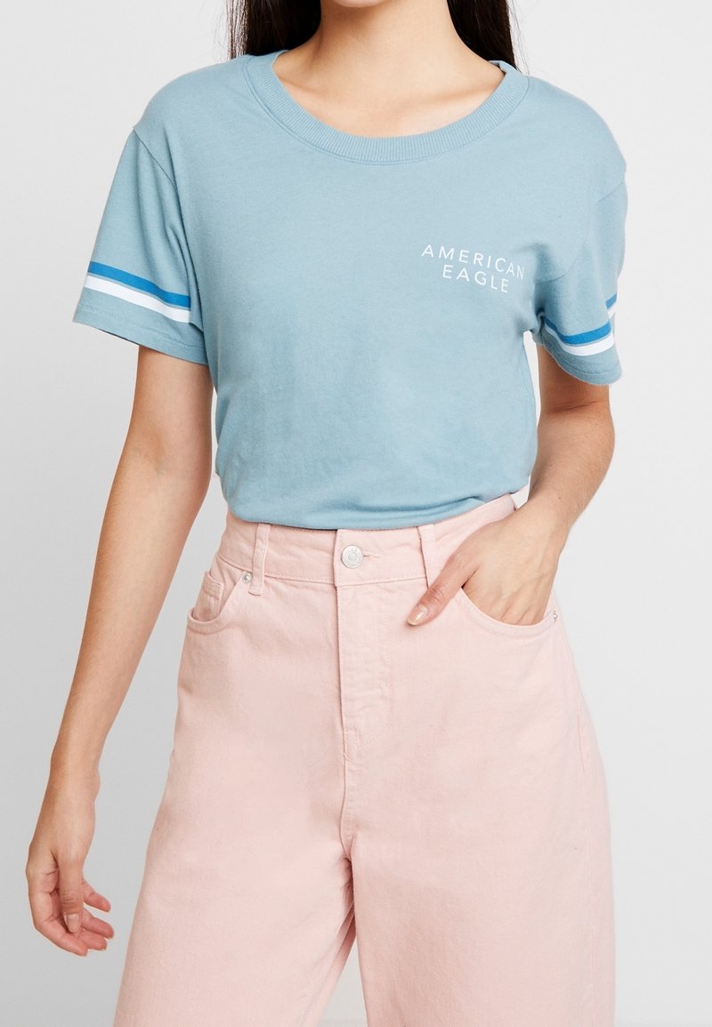 Femme portant un t-shirt American Eagle bleu clair avec des rayures blanches et bleues sur les manches et un pantalon rose clair taille haute.