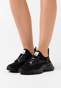 Baskets de sport noires en tricot avec un dessus en maille, dotées d'une semelle épaisse, de lacets blancs contrastants et d'une languette à l'arrière.