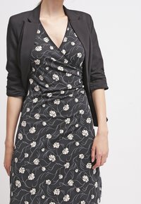 Zwarte blazer over een donkere bloemen wikkeljurk met crèmekleurige bloemen en vloeiende lijnen, gemaakt van zachte stof en met een v-hals ontwerp.