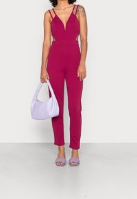 Tuta fucsia con una profonda scollatura a V, lati sagomati e spalline sottili. Abbinata a una borsa lavanda e sandali intrecciati lavanda.