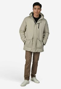Beige geïsoleerde parka met capuchon, ritssluiting aan de voorkant, twee grote voorzakken en zwarte ritssluitingdetails. Gedragen over een donker shirt en bruine broek.