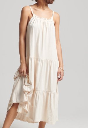 Femme portant une robe midi crème sans manches avec une jupe à étages et un délicat collier chaîne, tenant un tissu dans une main.