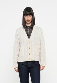 Cardigan in maglia beige con motivo a treccia, scollatura a V, orlo a costine, due tasche frontali e bottoni dorati. Indossato sopra una blusa nera a pois.