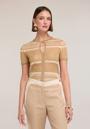 Kobieta o prostych brązowych włosach nosi beżową wzorzystą bluzkę z krótkim rękawem z dekoltem typu „keyhole” oraz beżowe spodnie z wysokim stanem i białymi bocznymi panelami.