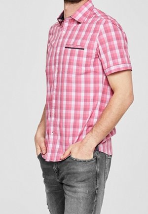 Homme portant une chemise à manches courtes à carreaux roses et blancs avec une poche poitrine à accents noirs, mains dans les poches, associée à un jean gris.