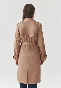 Beige Trenchcoat mit tailliertem Schnitt, Schulterpolstern und einem Rückenschlitz. Hat einen runden Kragen und ein rückseitiges Knopfdetail.