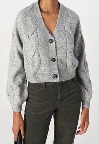 Grijze gebreide cardigan met een V-hals, vijf knopen en een gestructureerd kabelpatroon. Korte pasvorm met geribbelde zoom; gecombineerd met donkergroene corduroy broek.