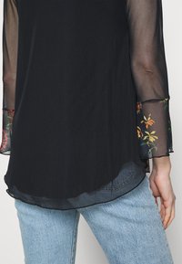 Sort langærmet top med gennemsigtige ærmer med blomstermønster og en buet kant, parret med lyseblå jeans. Blødt tekstureret stof.
