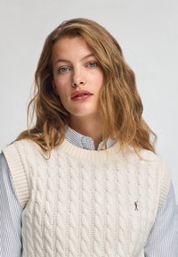Cremfarbener Kabelstrickpullover mit Rundhalsausschnitt, getragen über einem blau-weiß gestreiften Hemd. Verfügt über gerippte Armlöcher und ein kleines Logo.