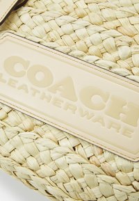 Wiklinowa torba ze gładką, kremową skórzaną metką z wytłoczonym tekstem "COACH LEATHERWARE". Tekstura jest naturalna i organiczna.