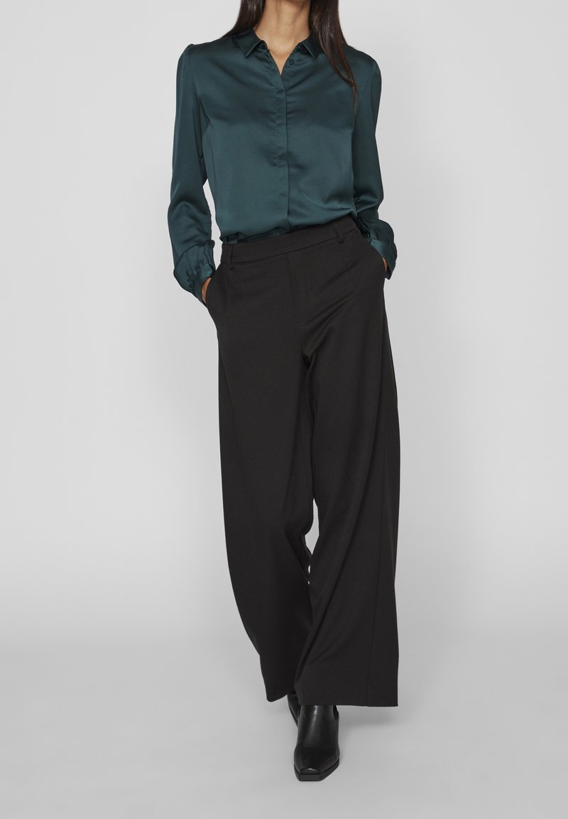 Femme portant un chemisier en satin turquoise rentré dans un pantalon noir taille haute à jambes larges, les mains dans les poches, chaussée de bottines noires.