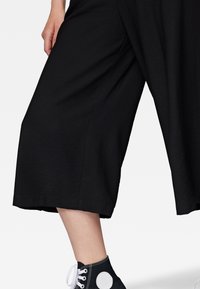 Black culotte pants with a loose fit and smooth texture, paired with high-top black sneakers featuring a white circular accent.
