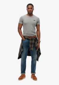 Grå kortærmet t-shirt, blå jeans, ternet skjorte knyttet om taljen, brun læderbælte og sko, med et ur på håndleddet.