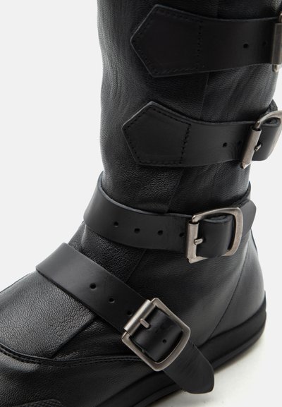 Μαύρο δερμάτινο μποτάκι μέχρι το μέσο της γάμπας με τρεις ρυθμιζόμενους ιμάντες και ασημένιες μεταλλικές αγκράφες πάνω σε υφή υλικού.