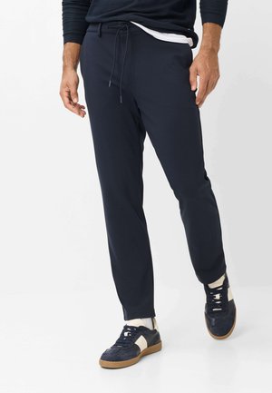 Mann trägt schwarze Jogger-Hose mit Kordelzug, weiße Socken und schwarz-weiße Sneakers, steht auf weißem Hintergrund.