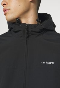 Veste zippée noire avec un col haut, des cordons de serrage réglables et un logo "Carhartt" brodé en blanc sur la poitrine. Texture de tissu lisse.