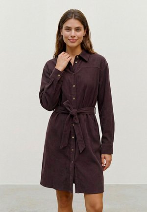 Une femme présente une robe-chemise à manches longues, de couleur prune foncé, avec des boutons dorés et une ceinture nouée à la taille, sur un fond clair et uni.