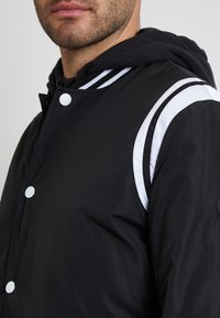 Schwarze leichte Jacke mit einem kontrastierenden weißen Streifendesign, Druckknöpfen und einer schwarzen Kapuze; glänzende Textur und sportlicher Stil.