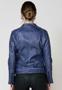 Cappotto in pelle blu con un design strutturato, dettagli cuciti e polsini con zip. Indossato sopra pantaloni neri, visto da dietro.