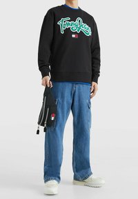 Felpa nera con "Tommy Jeans" ricamato in verde, abbinata a jeans blu larghi e sneaker bianche chunky. Una borsa nera è tenuta in mano.