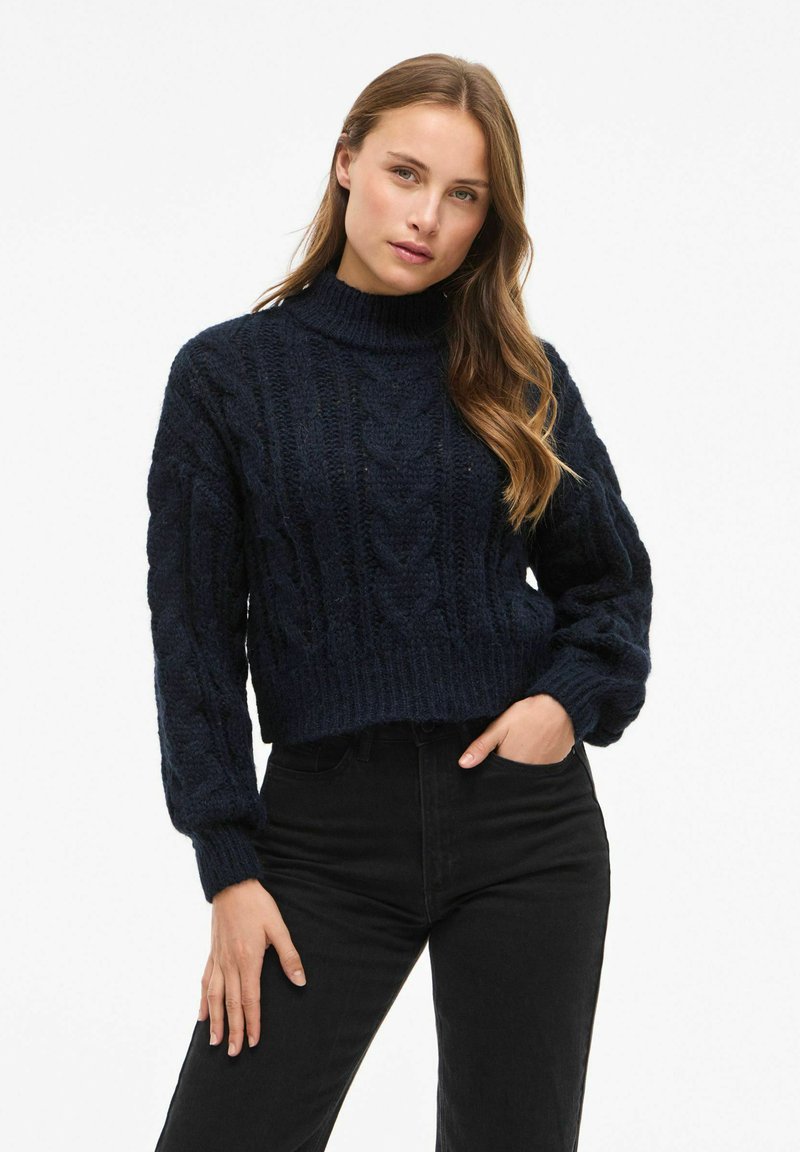 Granatowy, krótkie swetter z warkoczowym splotem, z wysokim golfem i ściągaczami, z wyczuwalnym wzorem i luźnym krojem. W połączeniu z czarnymi dżinsami.