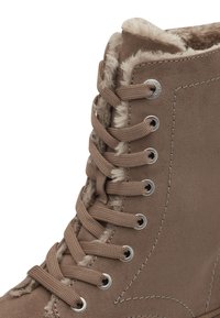 Botte lacée beige en suède avec une bordure en fausse fourrure en haut, œillets en métal et détails texturés sur le côté.
