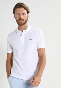 Polo bianco in cotone, con colletto classico, maniche corte e un logo ricamato verde sul petto.