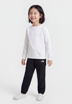 Enfant souriant portant une chemise blanche à manches longues, un pantalon cargo noir avec le logo The North Face, et des chaussures blanches à enfiler, debout sur un fond uni.