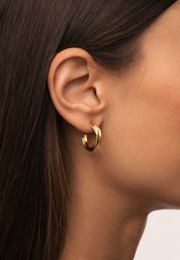 Boucle d'oreille en or avec un design torsadé, finition polie, et une épaisseur moyenne, portée sur l'oreille d'un modèle avec des cheveux raides.