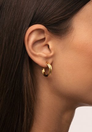 Orecchino a cerchio in oro con design intrecciato, finitura lucida e spessore medio, indossato all'orecchio di un modello con capelli lisci.
