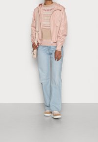 Ljusrosa nylonbomberjacka över en beige mönstrad tröja, tillsammans med ljusblå utsvängda jeans och bruna sneakers med vita sulor.