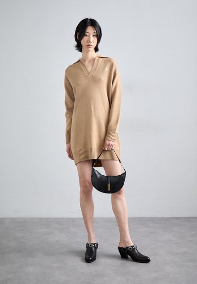 Beige gebreide jurk met een V-hals en een geribbelde zoom. Zwarte halfmaancycle tas met een gouden sluiting. Zwarte hoge schoenen met gespaccenten.