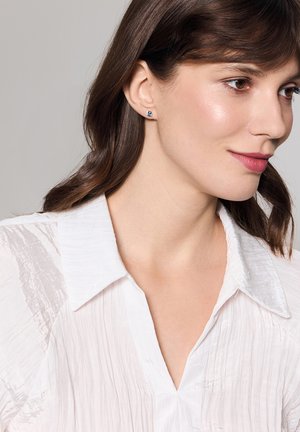 Okrągłe kolczyki z niebieskim kamieniem osadzonym w srebrze. Modelka nosi białą, fakturowaną koszulę z guzikami, prezentując naturalny makijaż.