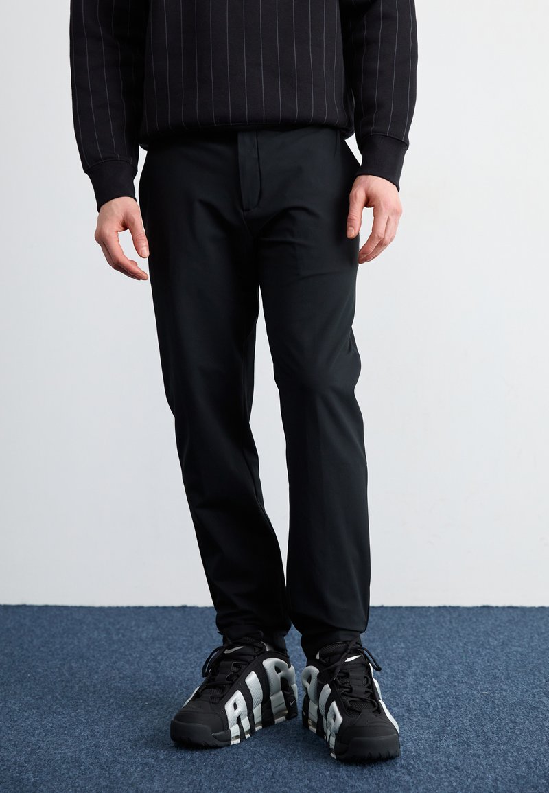 Sorte, slim-fit bukser med en glat tekstur, parret med kraftige sorte sneakers med fed hvid skrift og et robust såledesign.
