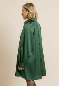 Robe en satin vert avec un col haut, des manches bouffantes et une coupe ample. Elle présente un dos plissé et des manches à poignets. Texture douce.