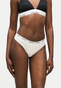 Γυναίκα που φοράει μαύρο bralette Calvin Klein και λευκά εσώρουχα Calvin Klein, στέκεται με τα χέρια χαλαρά στο πλάι μπροστά σε απλό φόντο.